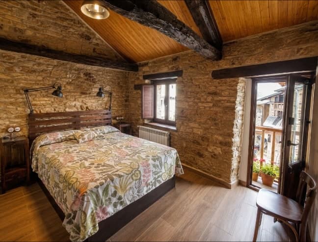 Suite Cereza: cama de matrimonio bajo el techo de vigas, muro de piedra y balcón sobre el Camino