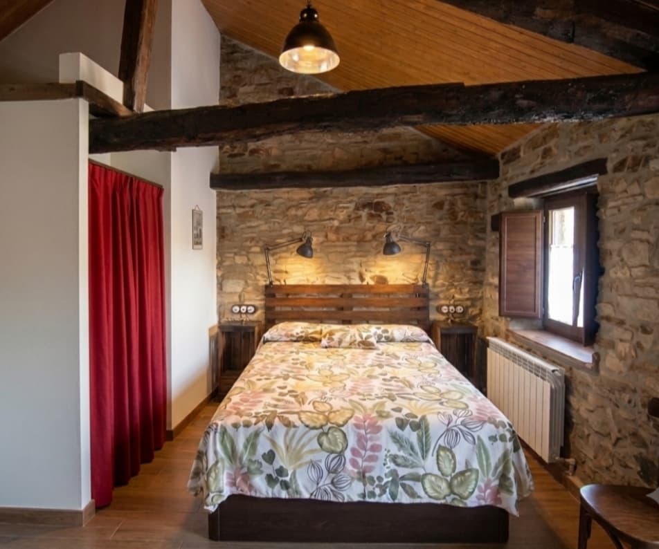 Suite Cereza: cama de matrimonio bajo las vigas, con muro de piedra y cortina roja